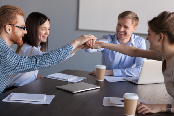 An image of a group of smiling workers around a table each putting their right hand into a stack as illustration of cooperation for post 'Employee Ownership: A Key to Staff Retention and Business Success?.'