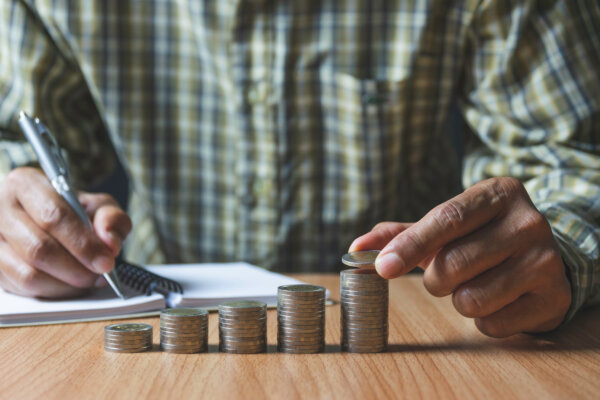 An image of a business owner sitting behind five piles of coins, increasing in height from left to right, with a notepad and pen, as illustration for post 'National Minimum Wage Increases - April 2025'