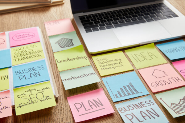 An image of a Mac Book and a range of coloured post-its laid out on a wooden desk with planning related terms written on them as illustration for post 'The pitfalls to avoid in Strategic Planning for 2025'